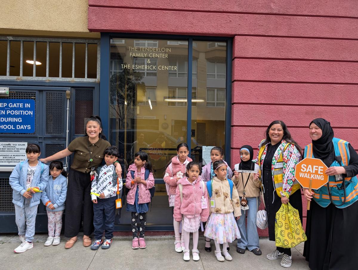 Students, caregivers and staff stand against a building and smile for a photo after completing a Walking School Bus.