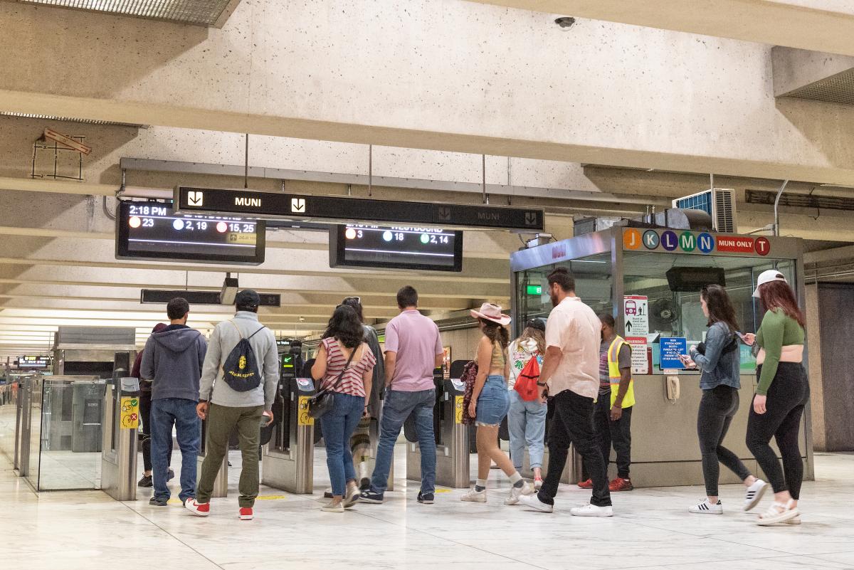 Several people enter fare gates at Muni Metro with digital displays noting arrival times above.