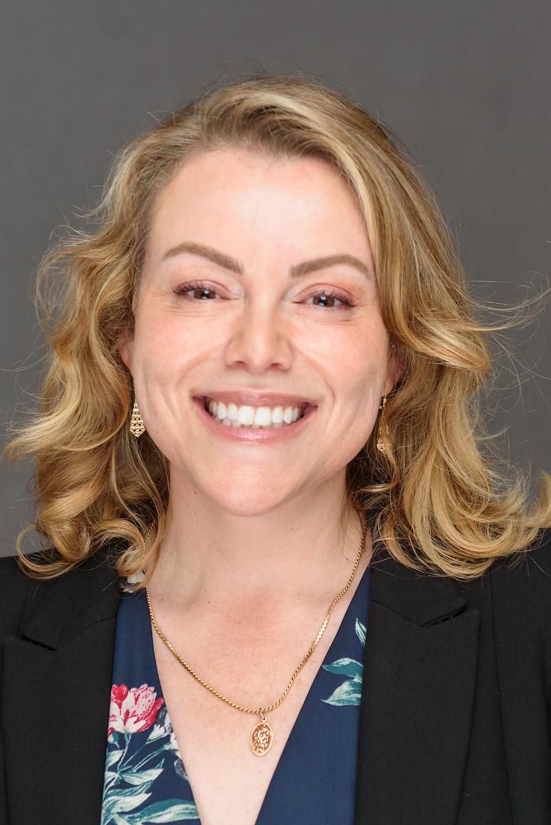 A person with light colored hair wearing a black blazer and blue patterned blouse.