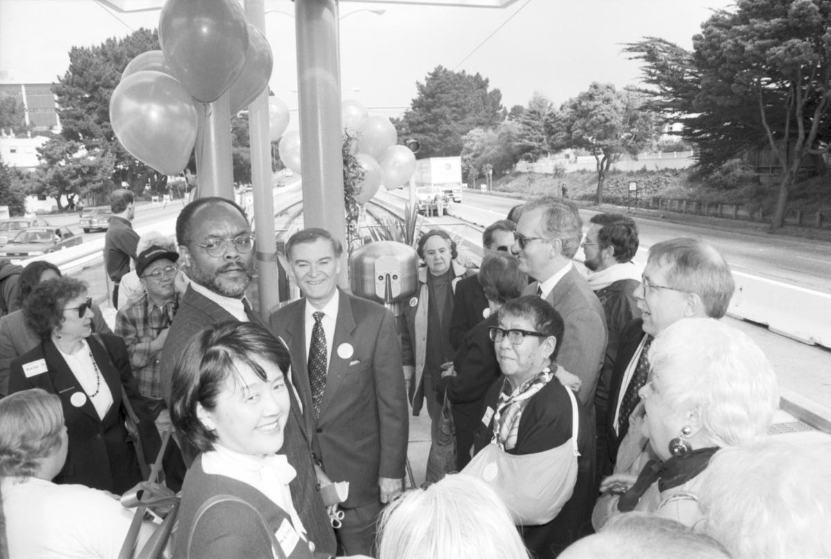 Group of people on an M platform by SF State posing for a ribbon-cutting. Photo includes Mayor Frank Jordan and Muni General Manager Philip Adams.