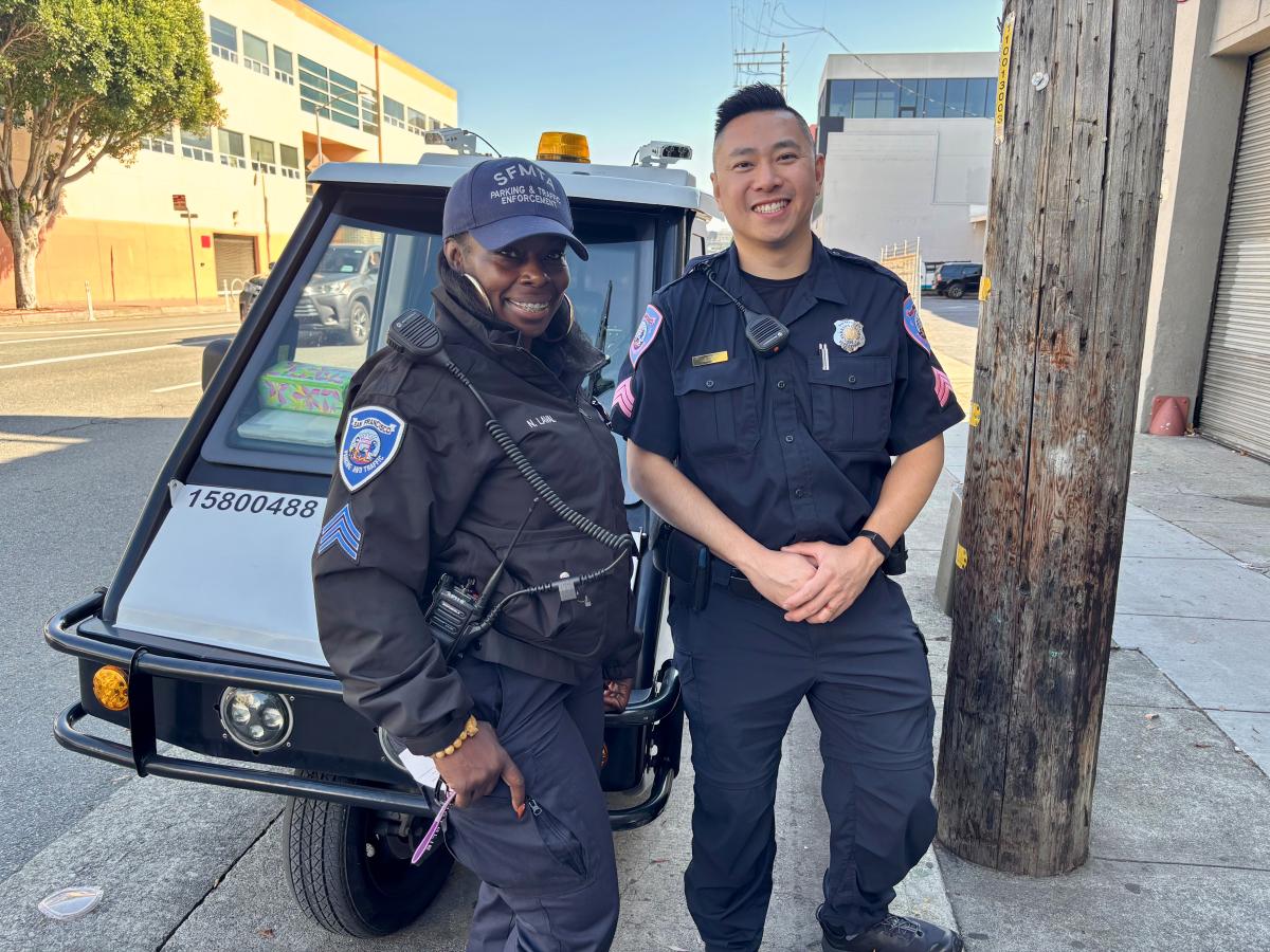 Two people in parking control uniforms stand in front of a city vehicle.