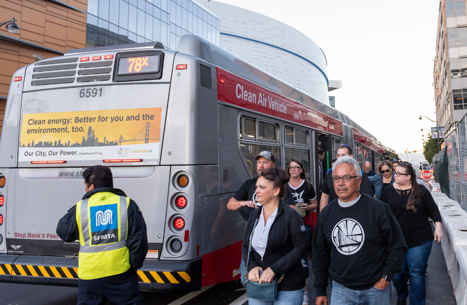 Several Muni riders exit a bus an SFMTA staffer in a yellow vest stands nearby.