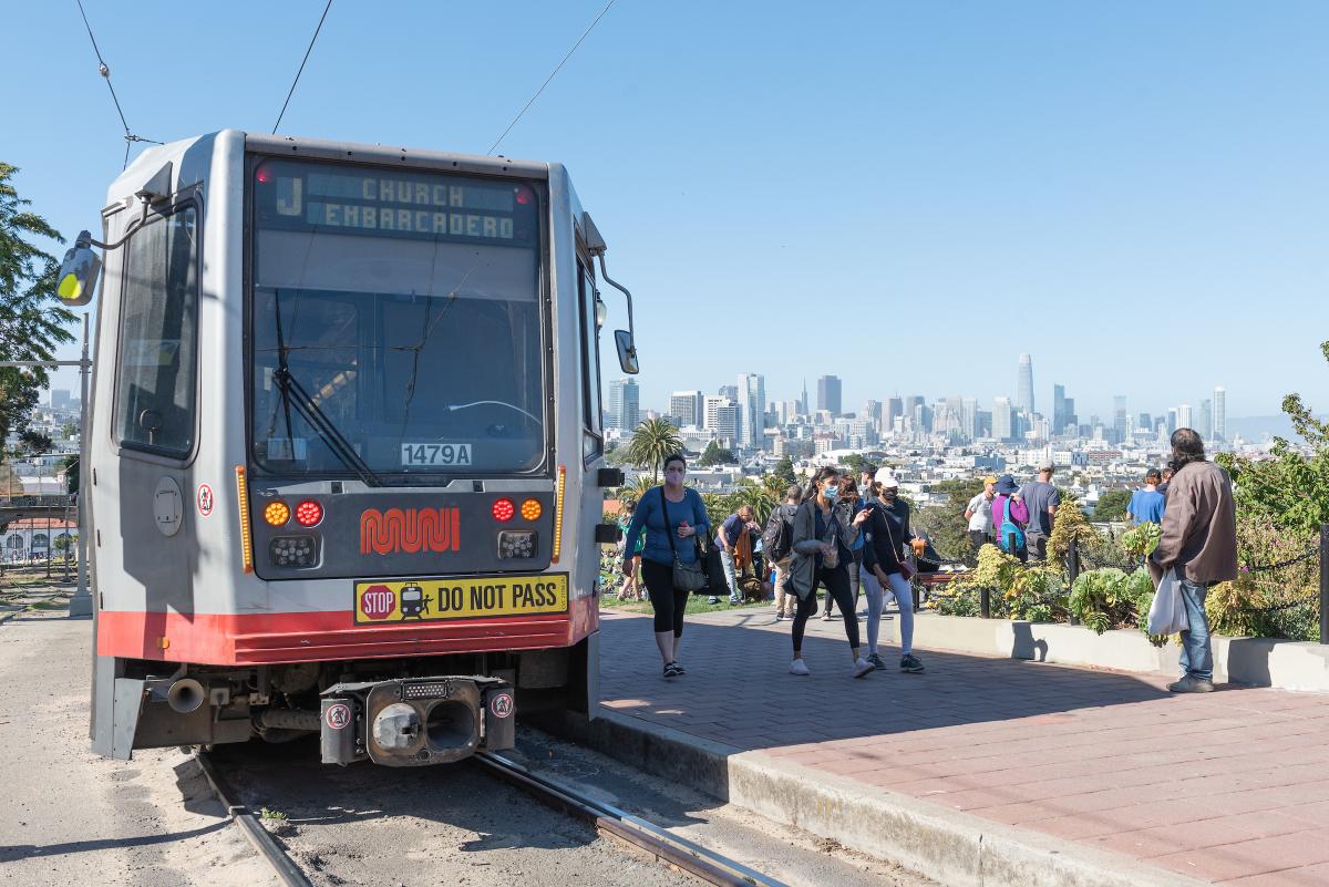 People near a train and light rail tracks with the skyline in the background.
