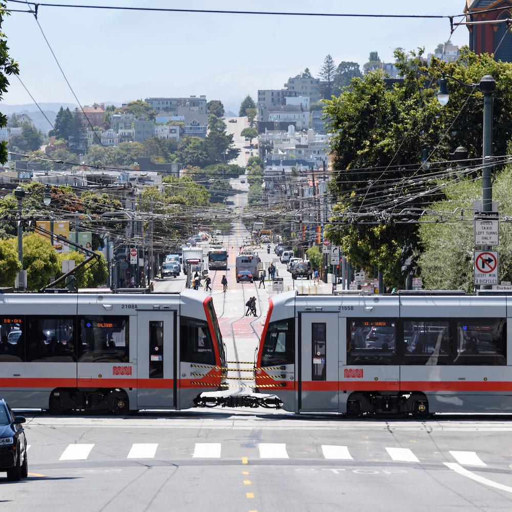 A light rail vehicle moves through an intersection with Muni buses in the background.