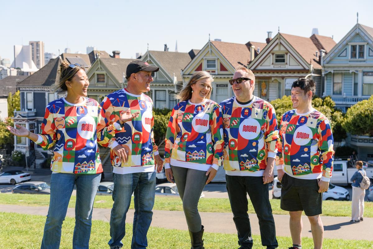 Several people smile in Patchwork of the City Holiday Pullovers as they stand in front of the Painted Ladies.