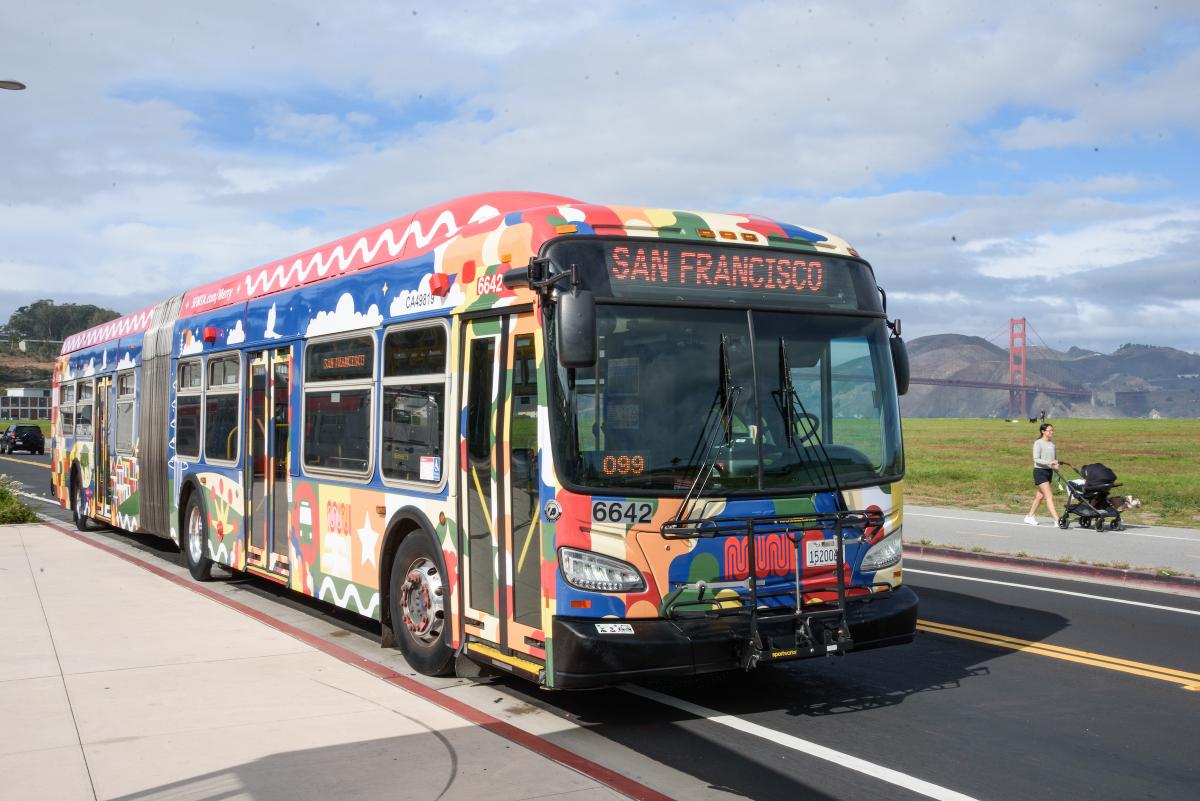 Bus wrapped with Merry Days of Muni artwork driving with Crissy Field and Golden Gate Bridge in the background.