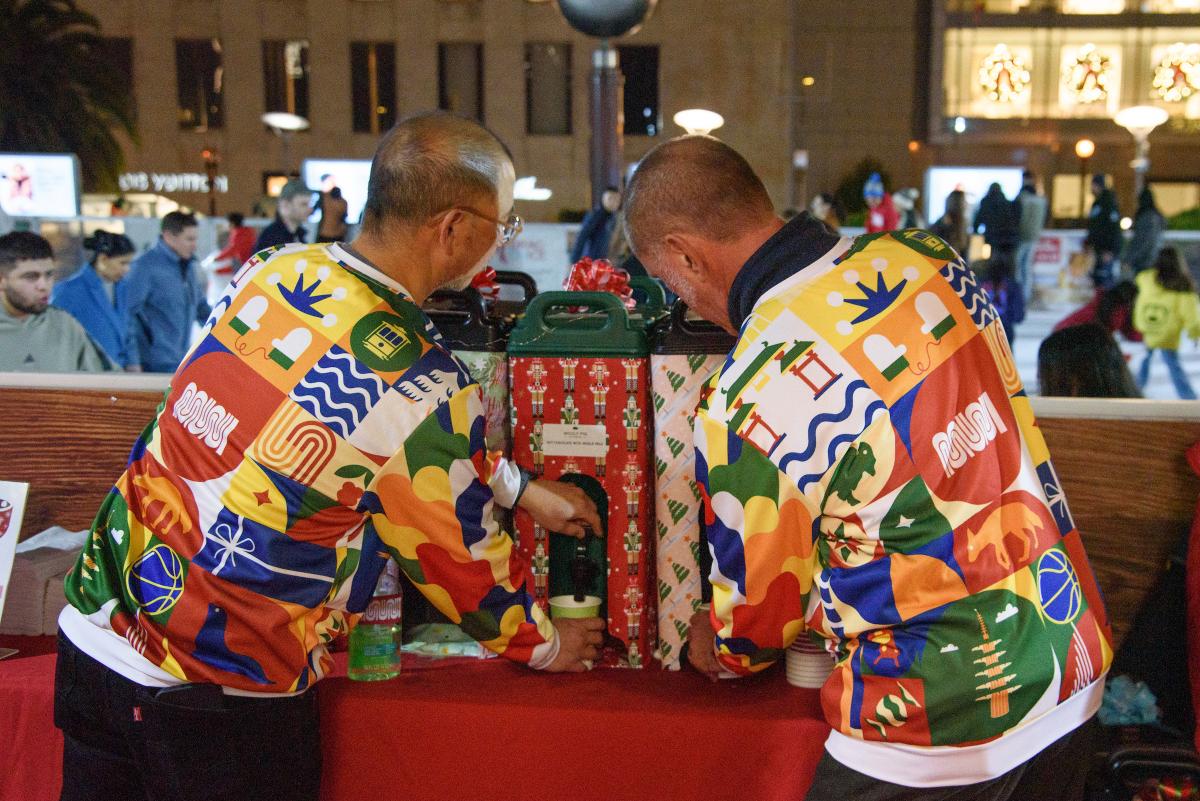 Two people wearing Muni holiday pullovers get hot chocolate for an event at the union square ice rink.