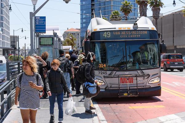 Several people board and exit a 49 Van Ness-Mission bus from a busy platform.