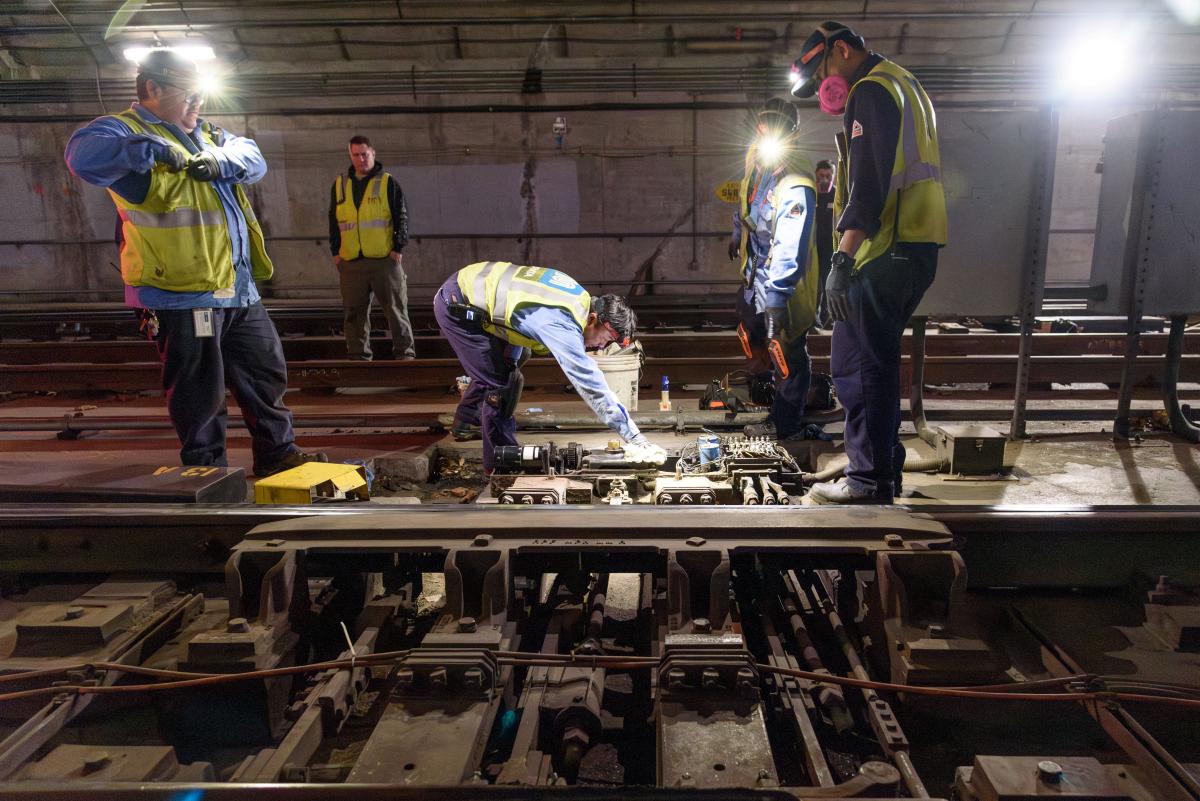 SFMTA crews wearing yellow vests complete work on rails in a Muni Metro tunnel.