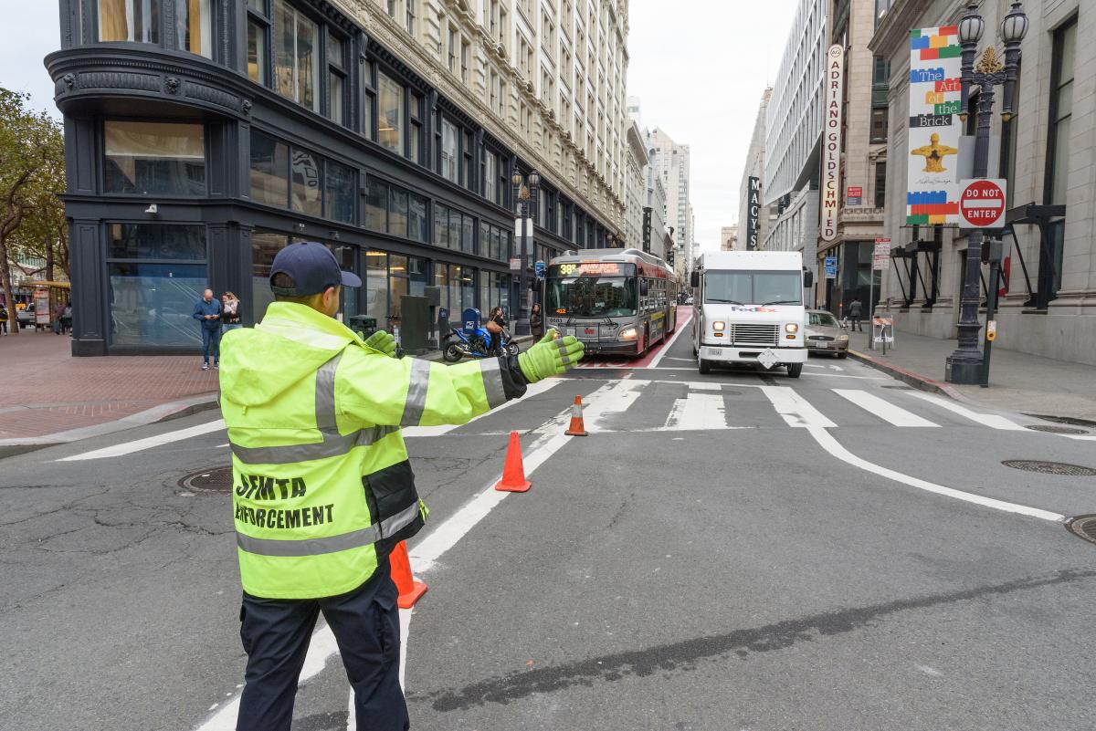 A parking control officer wearing a yellow safety jacket directs traffic near a downtown intersection.