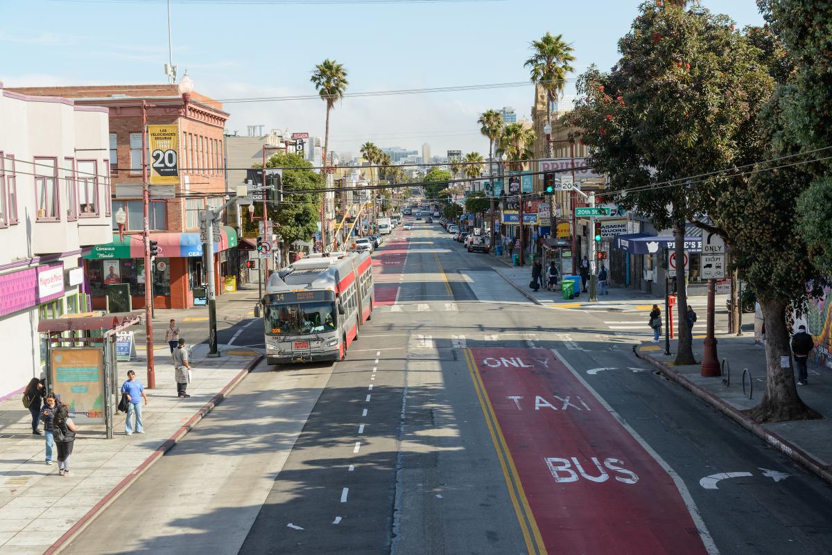 View of red transit lanes on Mission Street with a 14 Mission bus nearing a stop where people are waiting to board.