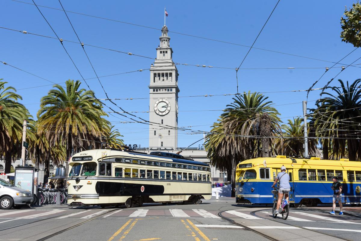 Two F Market historic streetcars turn onto Market with the Ferry Building behind them.