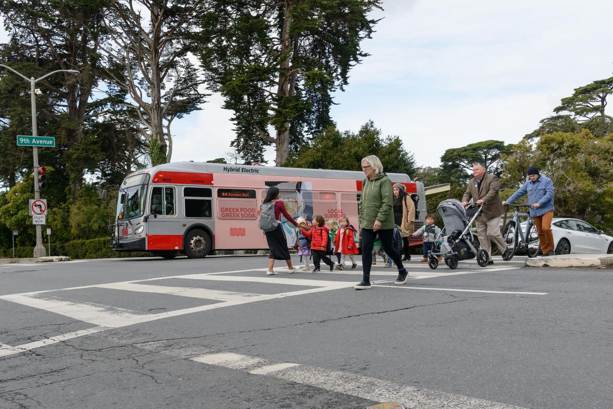 A bus crosses a street as several children and adults pushing strollers walk in an intersection.