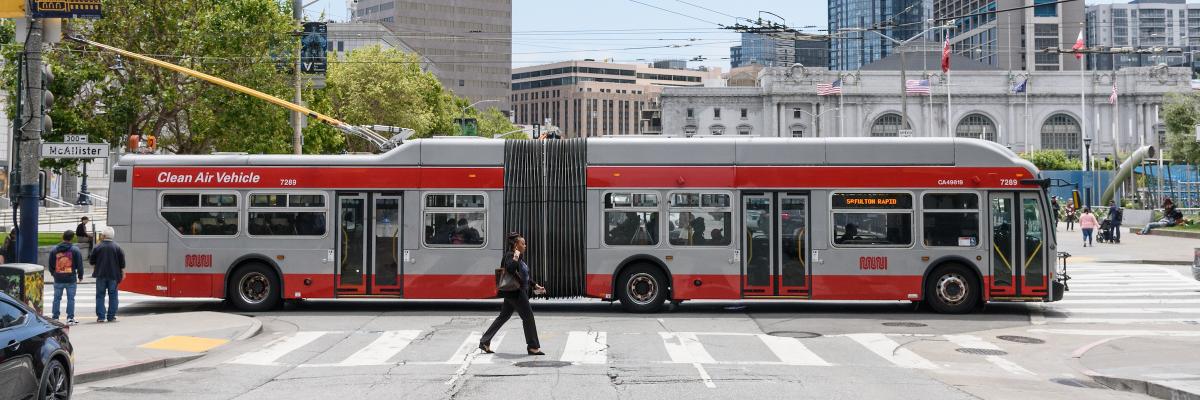 People cross a street by city hall as a 5 Fulton bus passes in the background.