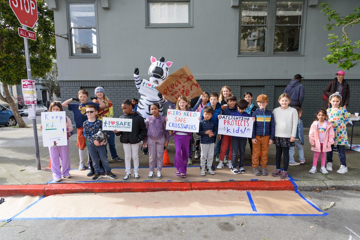 Several students hold signs with street safety messaging near a curb painted red to comply with state's daylighting law.