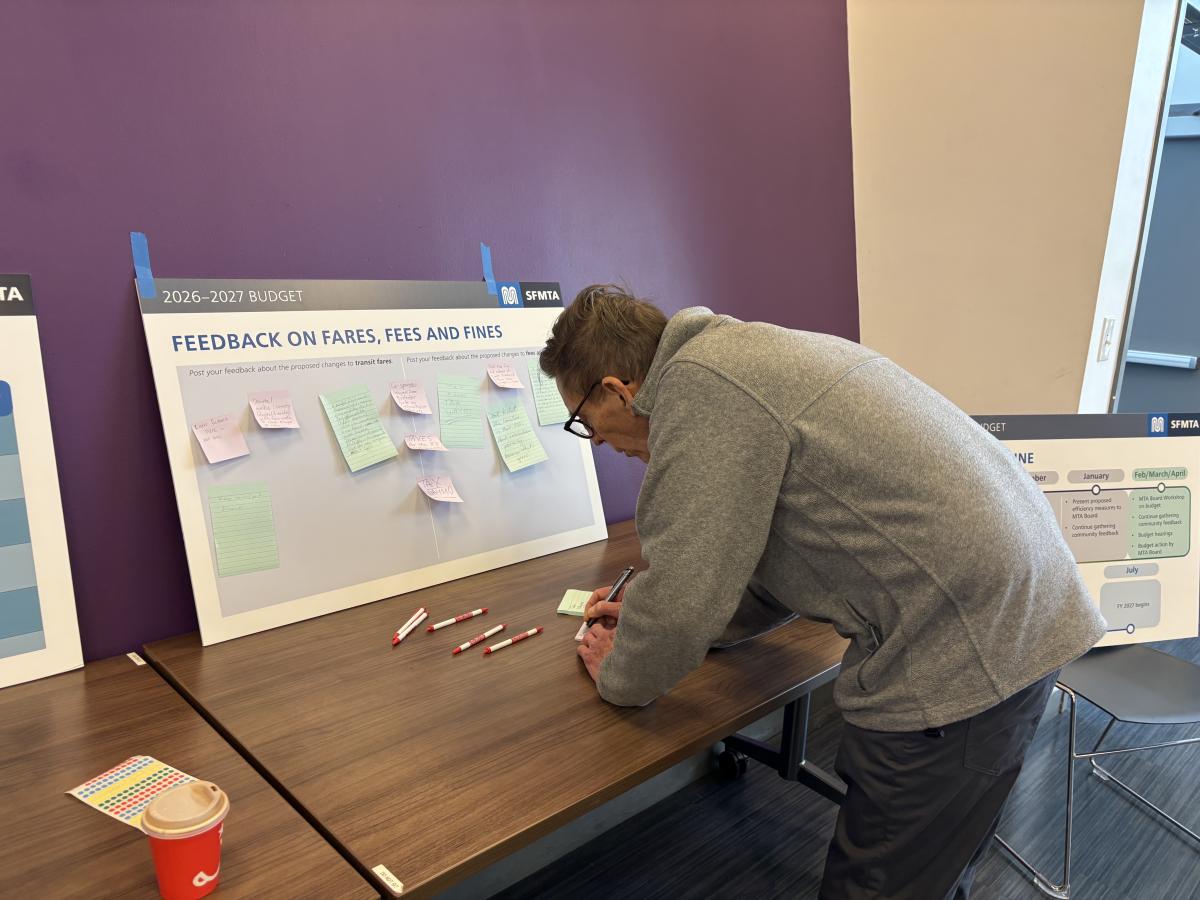 A person writes comments at a poster about feedback on fares, fees and fines at an SFMTA Budget Open House.