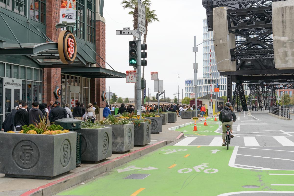 People walking on the sidewalk and a person riding in a green protected bike lane near Berry Street and 3rd Street in San Francisco. 