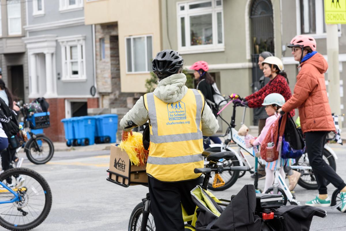Adults and kids riding bikes and scooters in front of buildings.