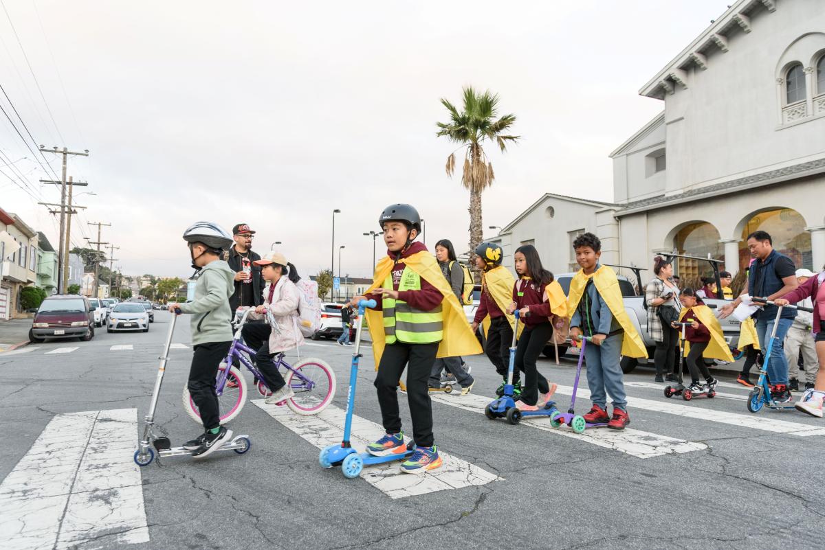 Adults and kids riding bikes and scooters through a crosswalk. Many students are wearing gold capes.