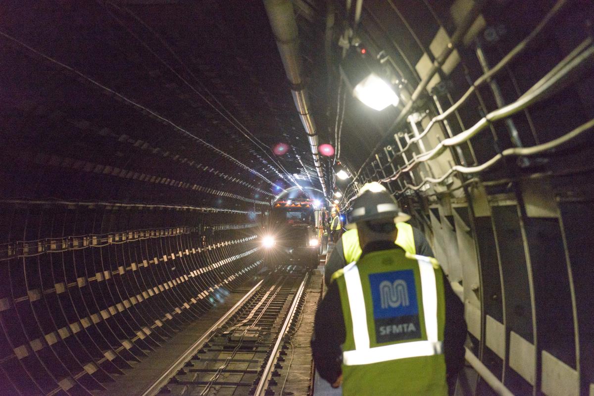 Two people wearing yellow safety vests walk in a Muni tunnel toward a vehicle on the tracks.