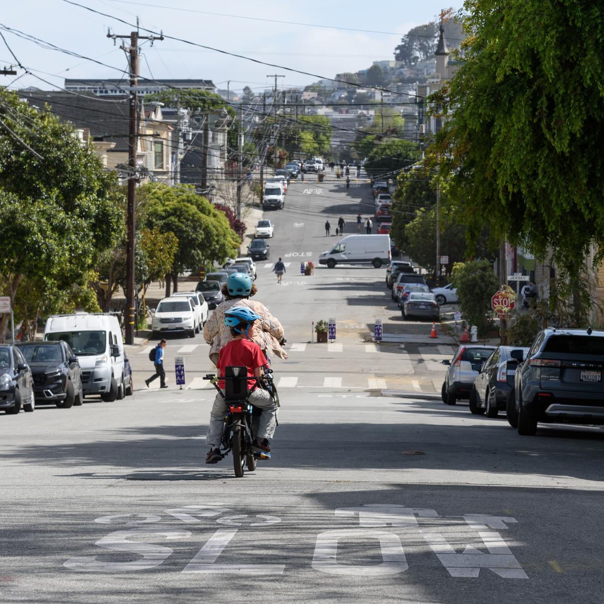 An adult and child ride a bike together down the street wearing helmets