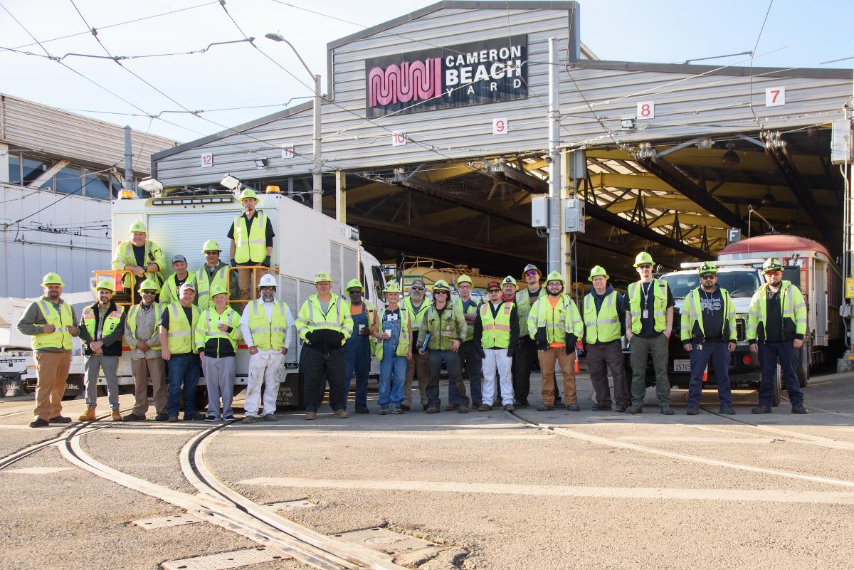 Several people wearing yellow safety vests stand for a photo in front of a building that says Cameron Beach Yard.