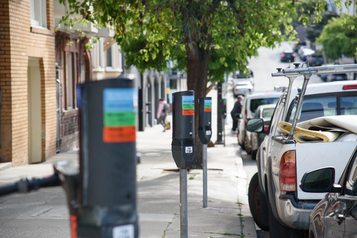 Closeup of SFMTA parking meters on a sidewalk near parked cars.