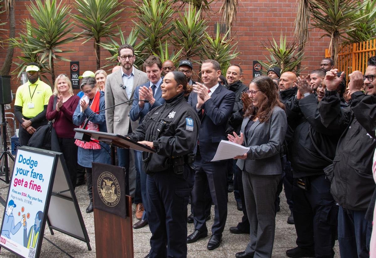 people gathered around a lectern as one person speaks to an unseen crowd. People are in various manners of dress, including dressed in formal wear as well as in transit enforcement uniforms