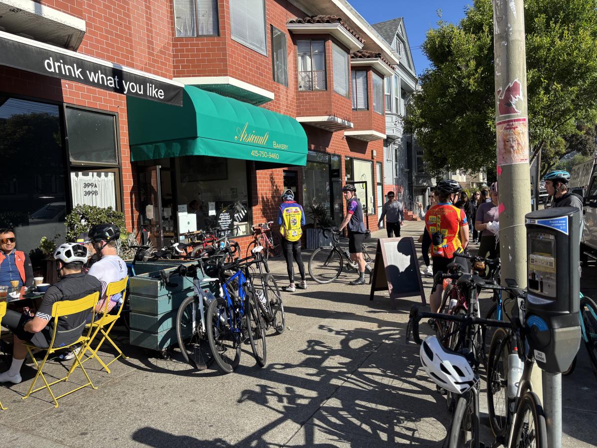 People sitting and standing near two cafes with bikes parked on the sidewalk.  
