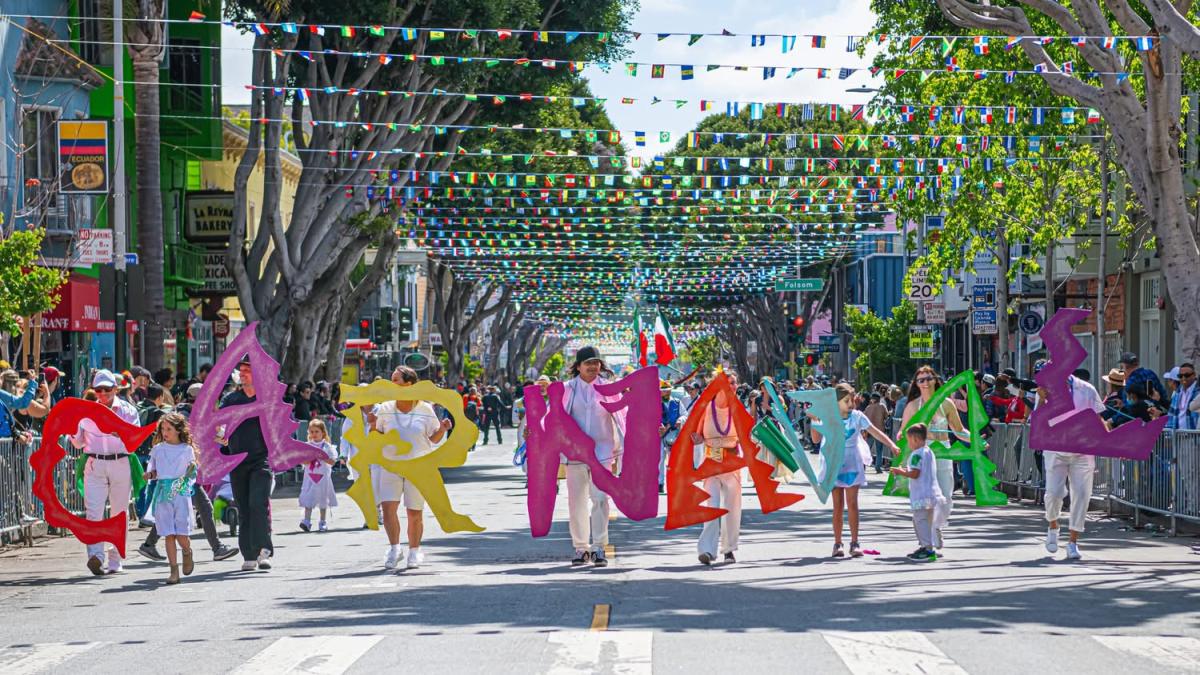  People marching in the street in a parade, holding up letters that spell Carnaval. 
