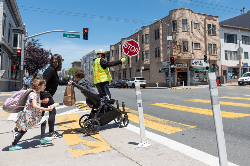 Adult with stroller, holding a child’s hand, getting ready to cross the street.