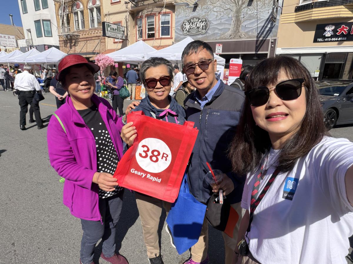 four people smiling and taking a selfie at a street fair. one holds up a red tote bag that says 38R Geary rapid