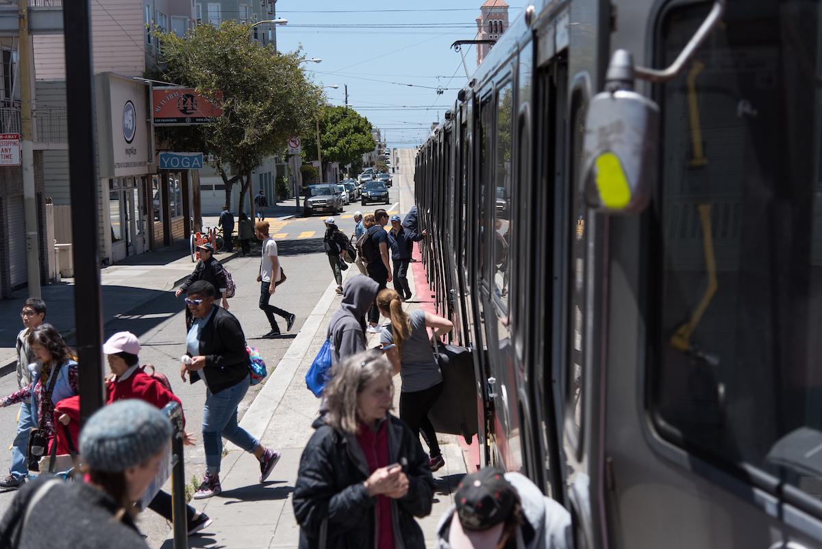 People board and walk near a Muni Metro train at a street-level stop in San Francisco on a sunny day. 