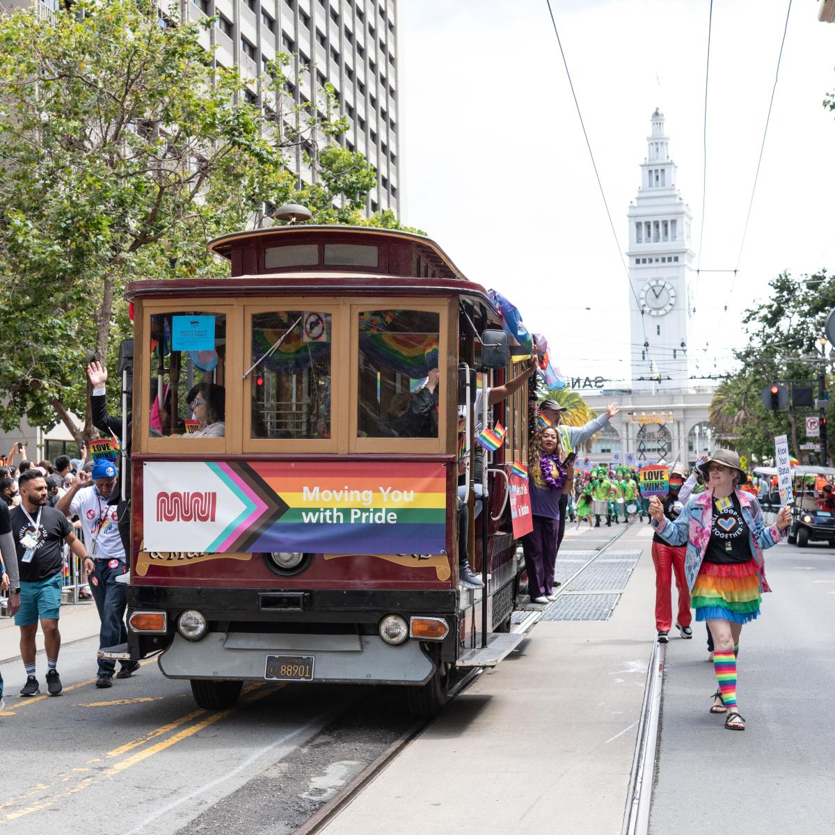 People dressed in colorful outfits march alongside a cable car. The cable car is decorated with a rainbow banner that reads "Moving you with pride"