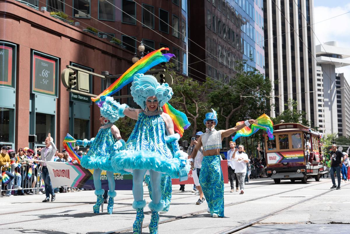 Three people dressed in neon blue outfits, hats and wigs wave pride flags and march down Market Street
