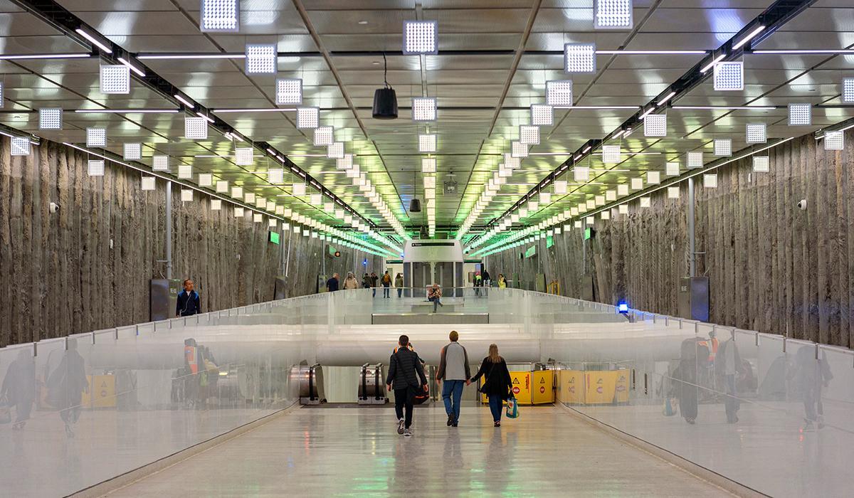 People walk through the Powell / Union Square–Market Street Station concourse beneath Lucy in the Sky, an illuminated ceiling artwork made of rows of square LED light panels. 