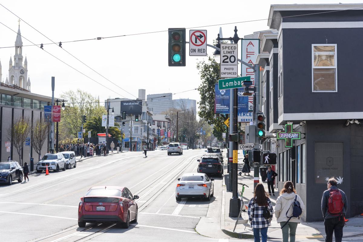 People walking and driving near a street corner that has a speed safety camera.