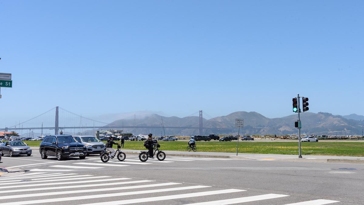 Cars and bikes at an intersection with grass behind it and a bridge in the background.     
