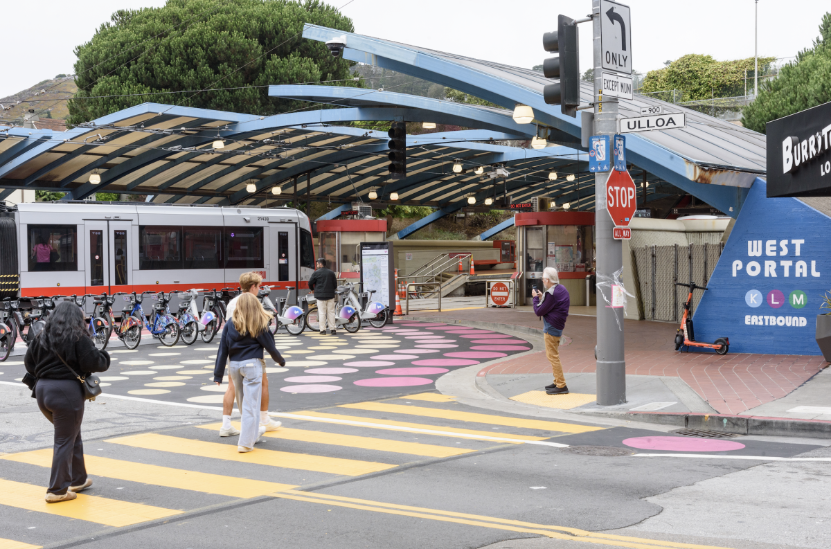 Outside of West Portal Station, several people cross the street near a street mural and bikeshare station while a train pulls in.