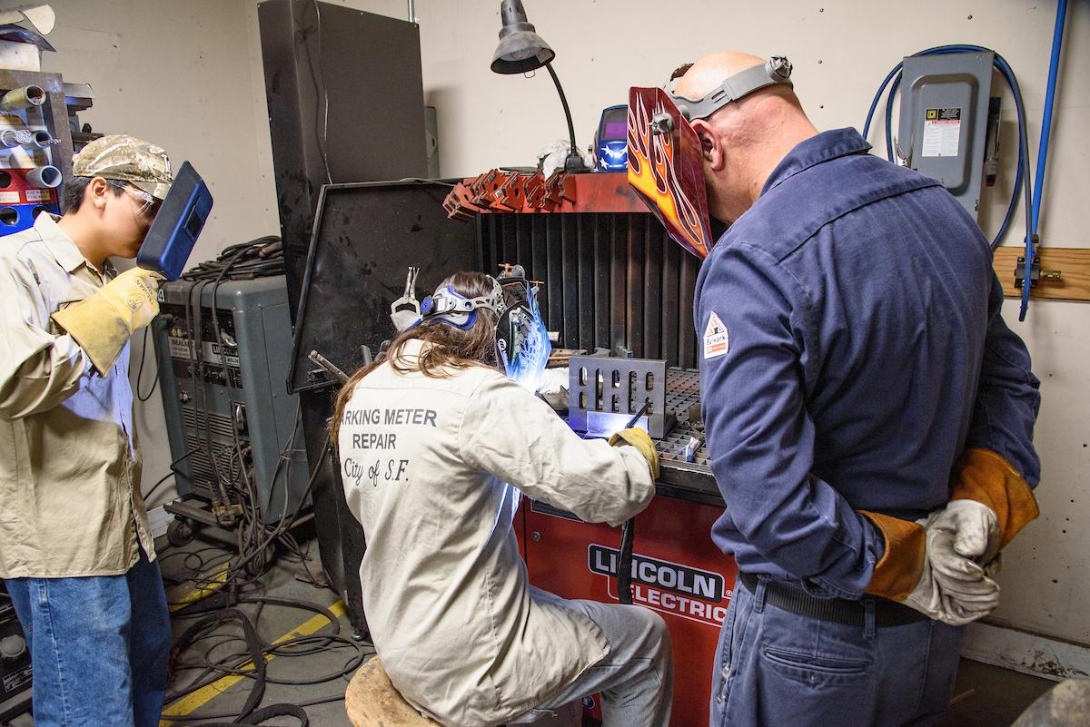 People in work uniforms wearing welders’ masks working on a piece of equipment.