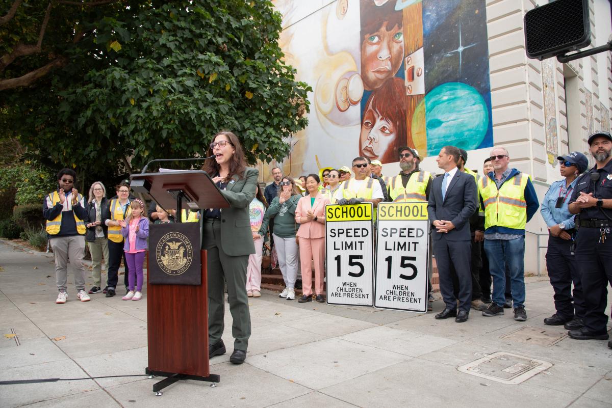 SFMTA Director of Transportation Julie Kirschbaum speaks at a podium. Staff hold speed limit signs behind her. 