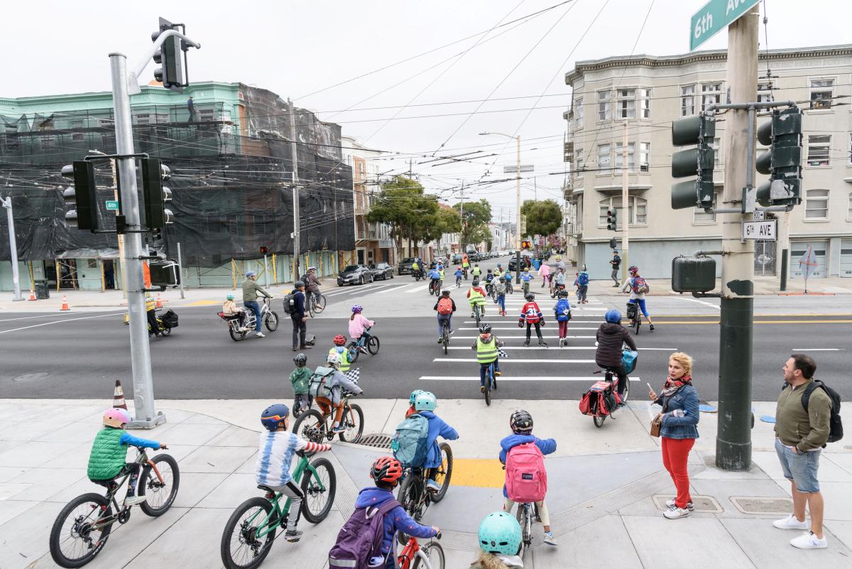 Students wearing helmets and safety vests bike across a street during a school event.