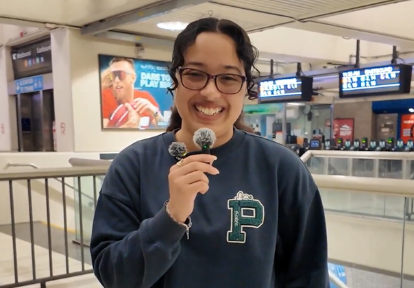  A person wearing a blue sweatshirt and holding a microphone in a transit station.  