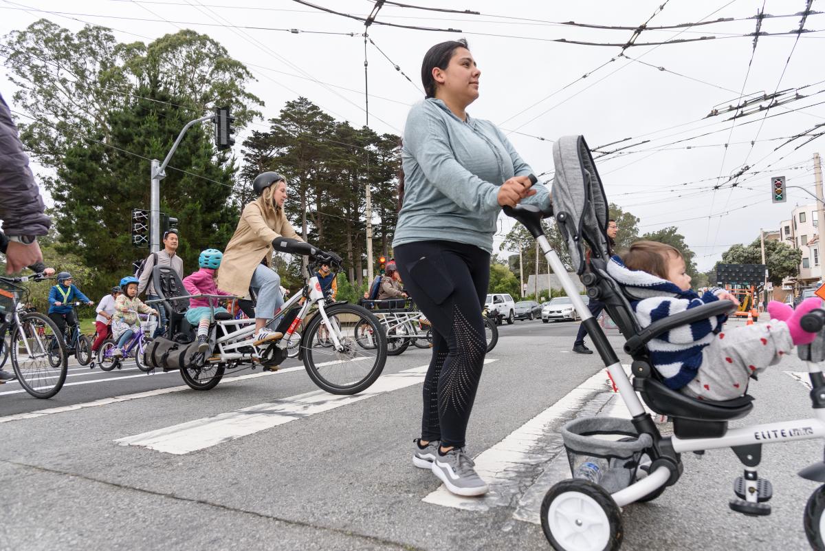 Adults and children ride bikes near people pushing strollers across a street during a school event.