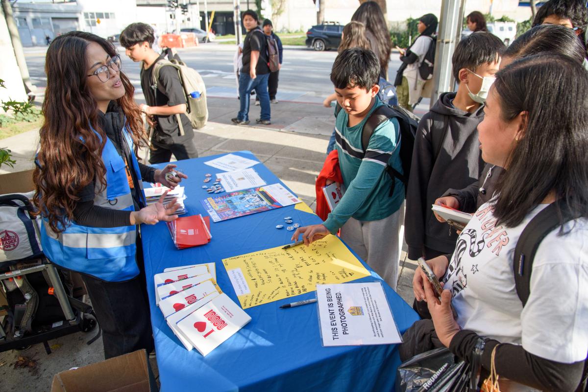 Students gather near a table where a staff member wearing a blue vest talks to them during a school event.
