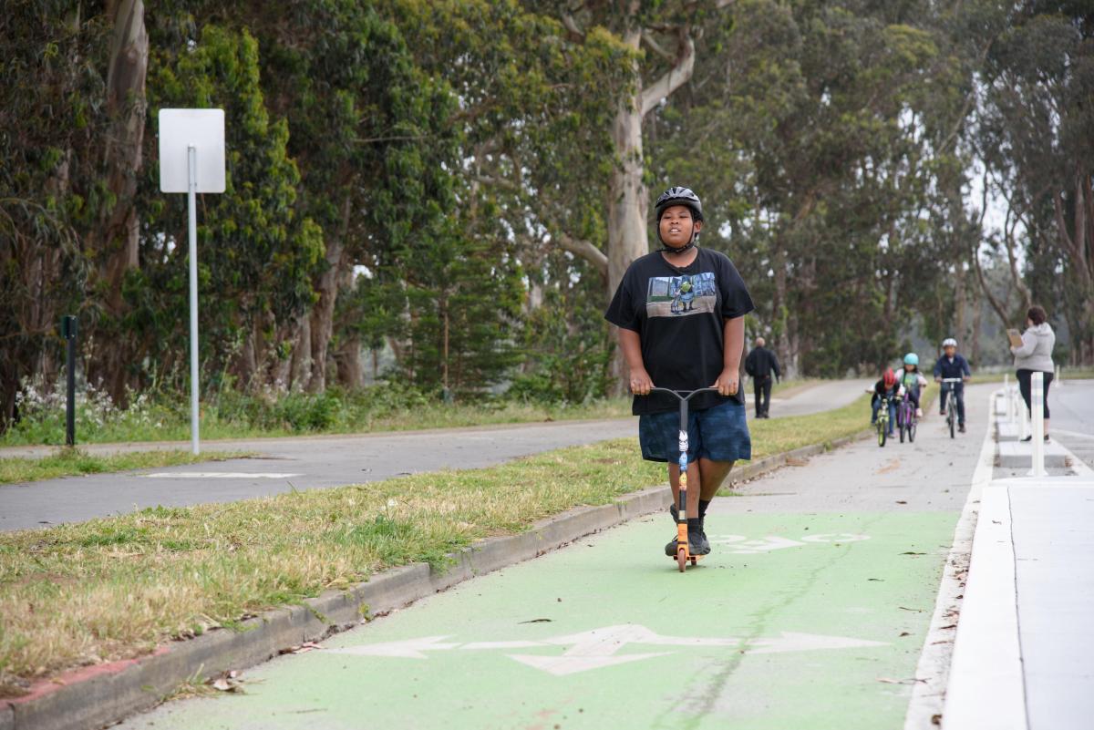 Una persona joven usa un scooter o patineta en una ciclovía verde separada del tráfico. 