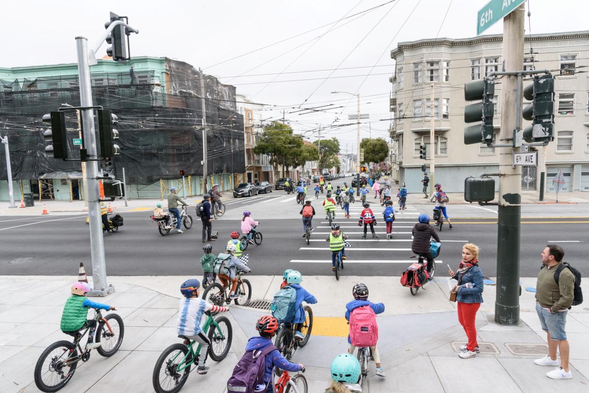 Estudiantes con cascos y chalecos de seguridad andan en bicicleta por una calle durante un evento escolar. 