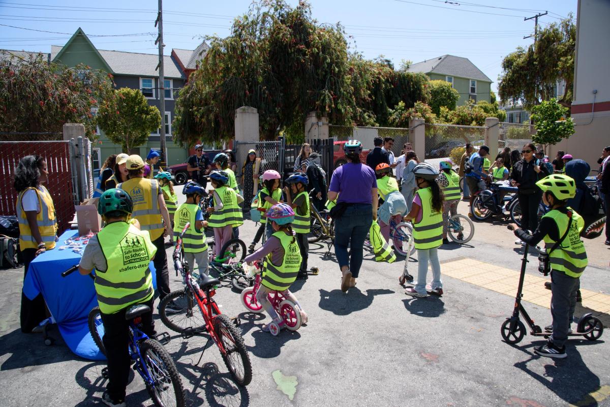 Estudiantes con chalecos amarillos paran con sus bicicletas cerca de una mesa con personal durante un evento escolar.    