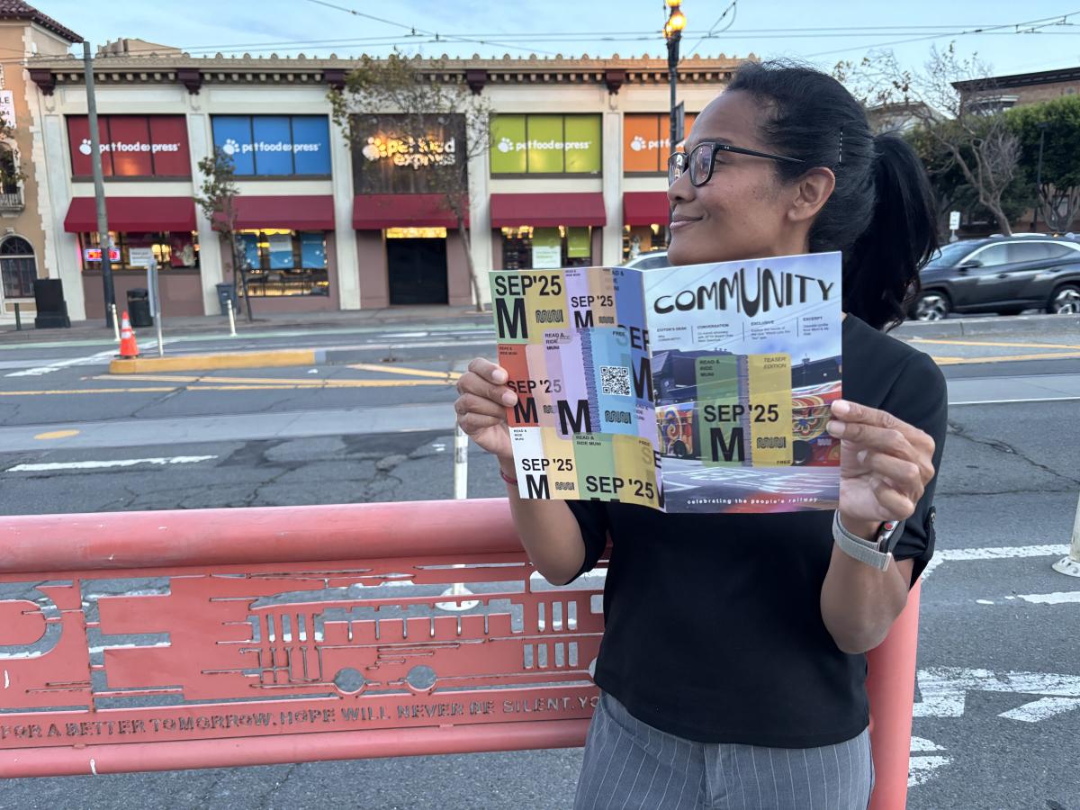 A woman standing near a street and holding a magazine. A colorful building is behind her. 