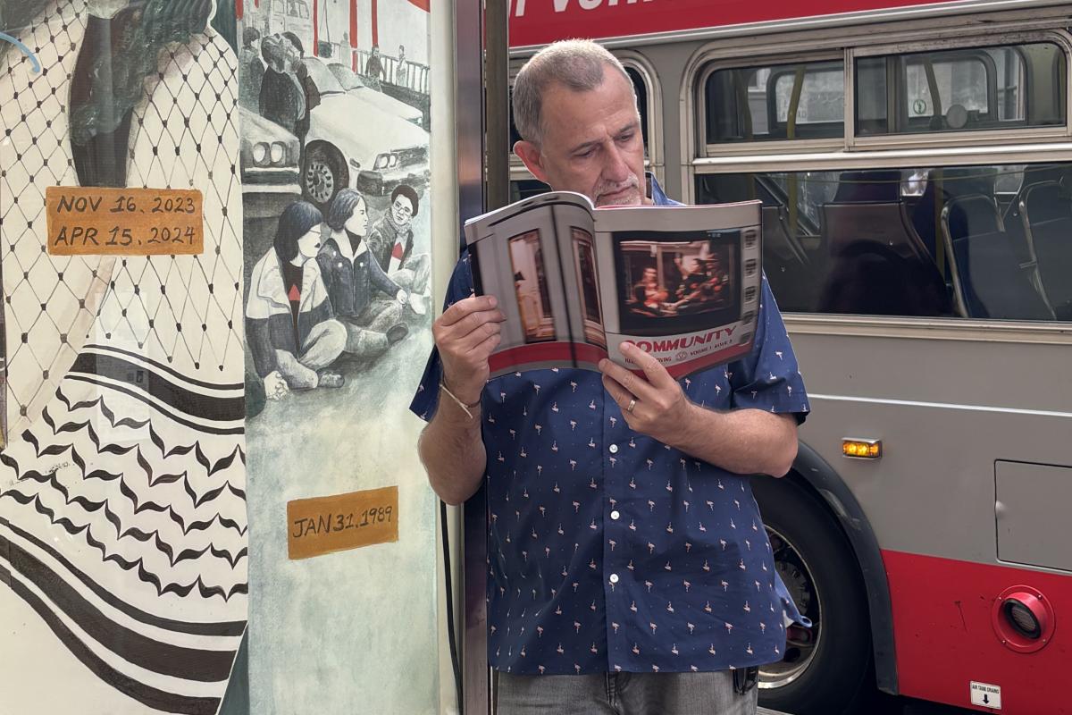 A man standing at a transit stop reading a magazine. A bus is behind him.
